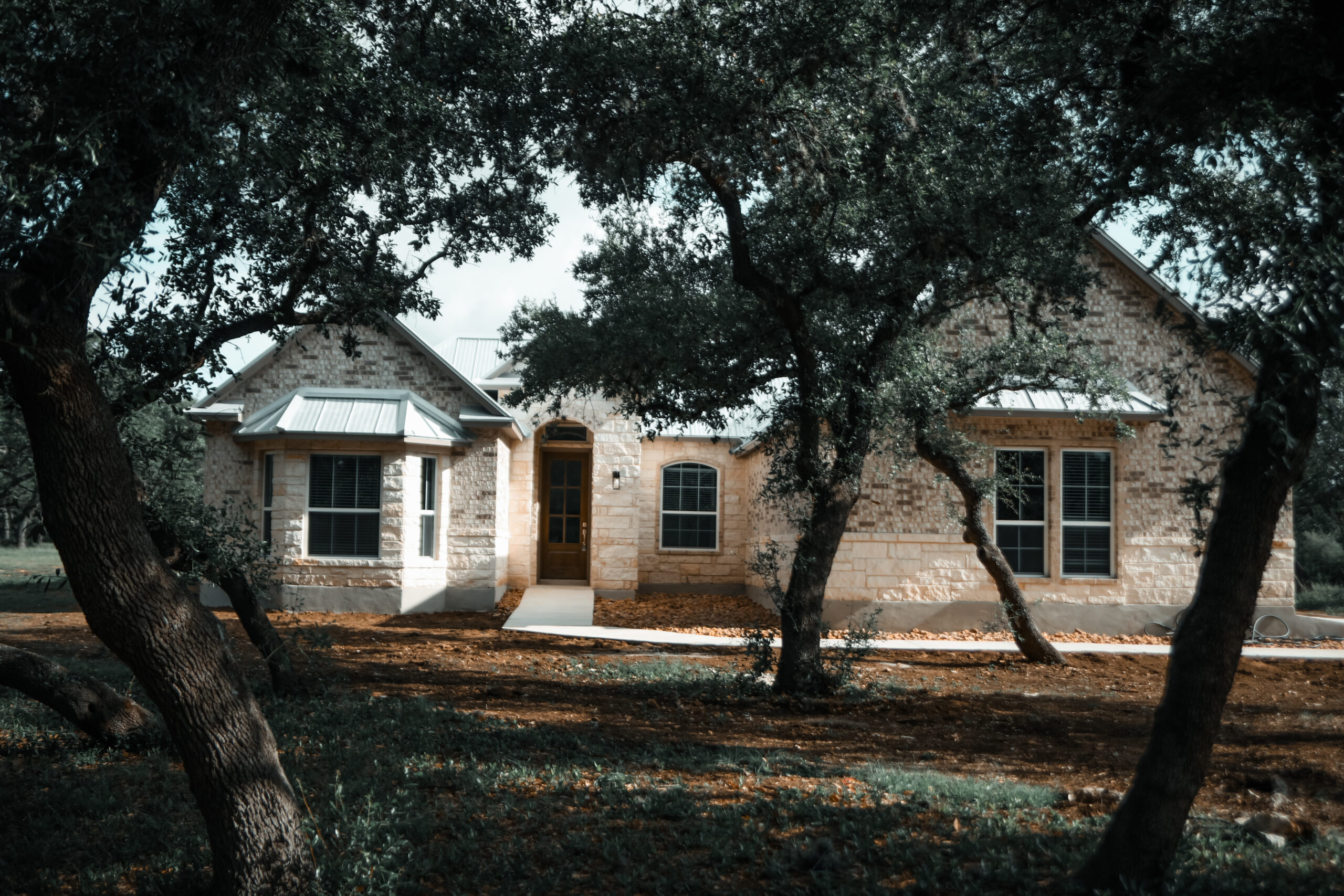 Single-story stone house with multiple large windows, a central wooden door, and a metal roof, partially shaded by several mature trees in the front yard.