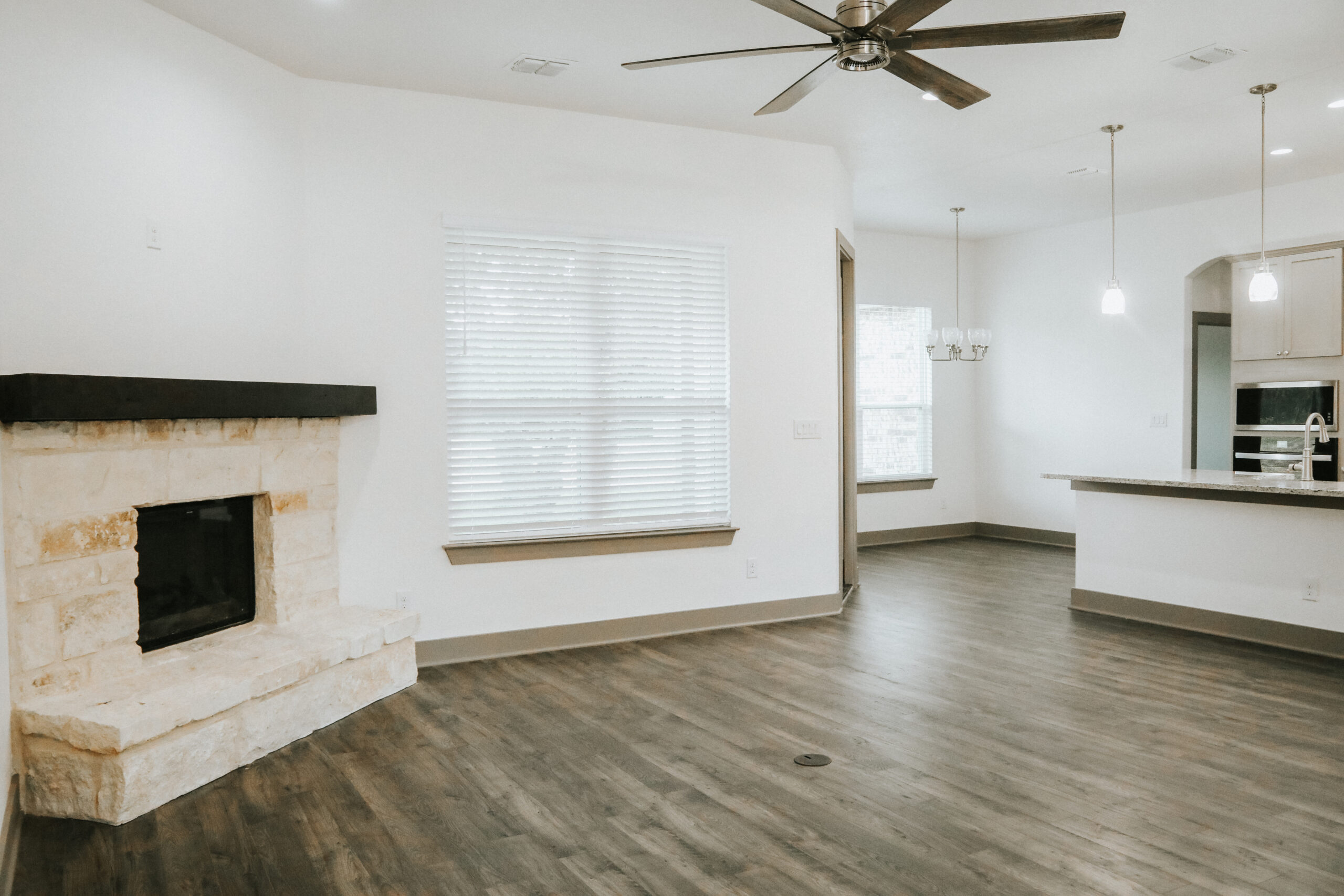 Empty modern living space with wood flooring, a stone fireplace, large window with blinds, ceiling fan, and open kitchen area with pendant lights.
