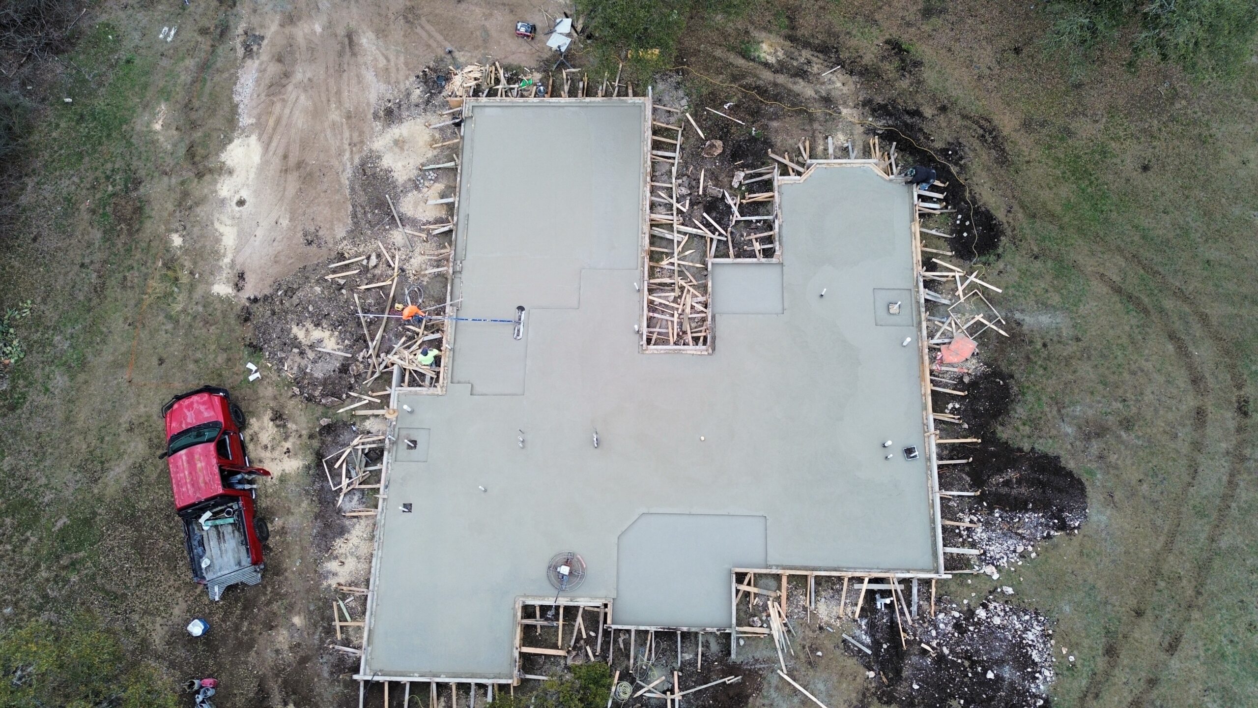 Aerial view of a freshly poured concrete foundation for a house, surrounded by wooden framing and construction materials, with a red truck parked nearby.