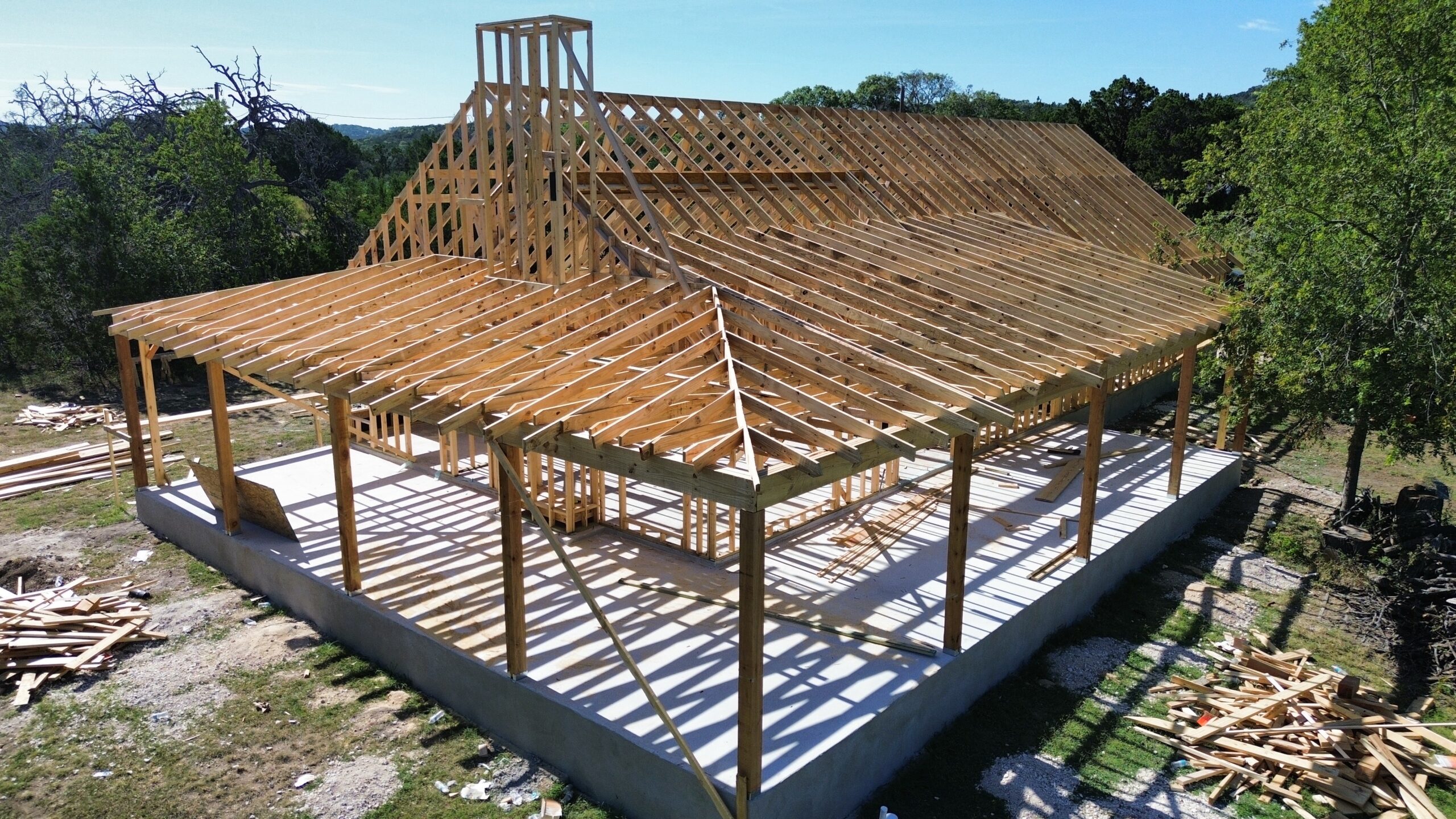 A partially constructed house with wooden framing and roof trusses on a concrete foundation, surrounded by trees and building materials.