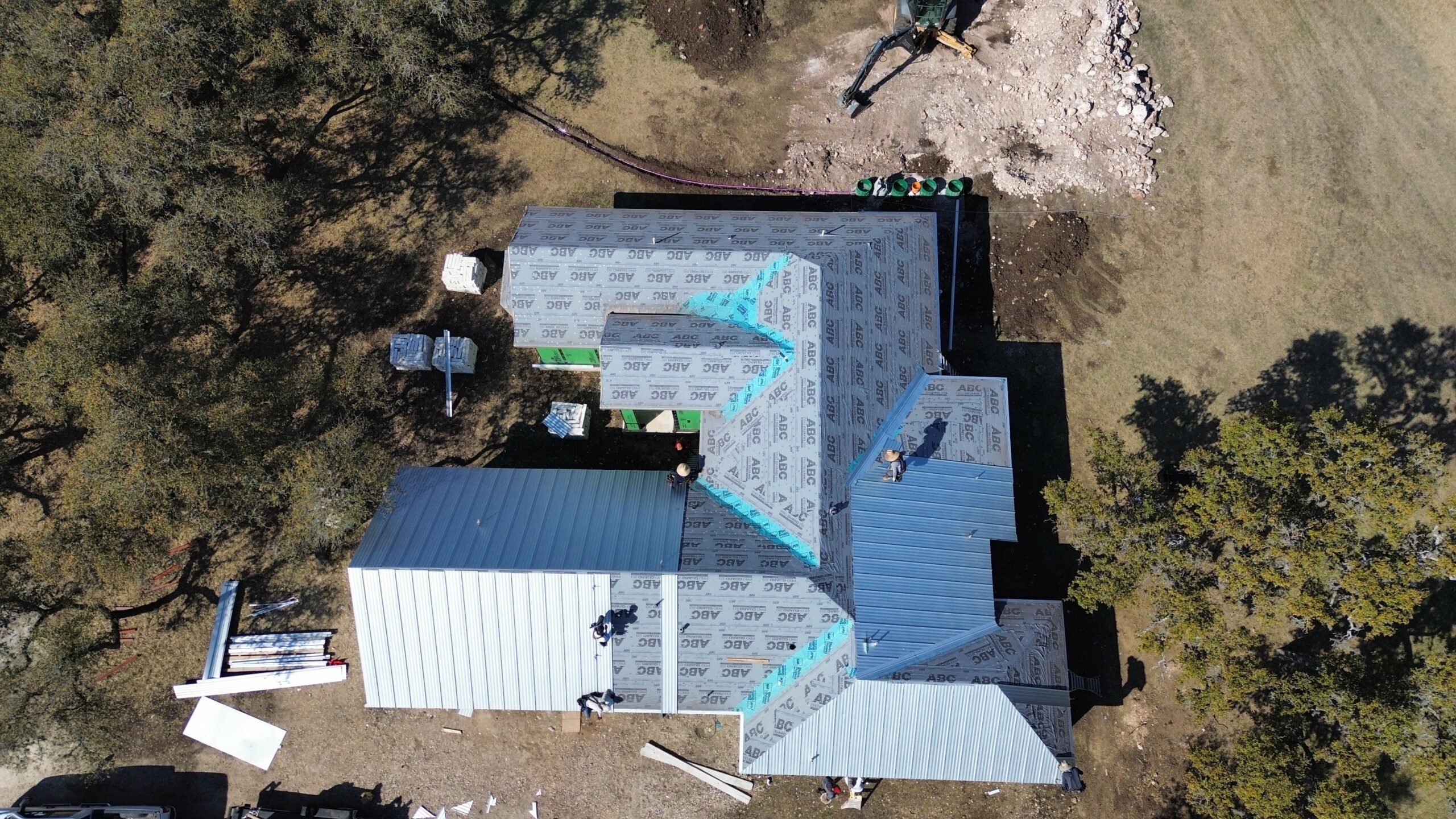 Aerial view of a house under construction, workers installing roofing materials, surrounded by trees and construction equipment.