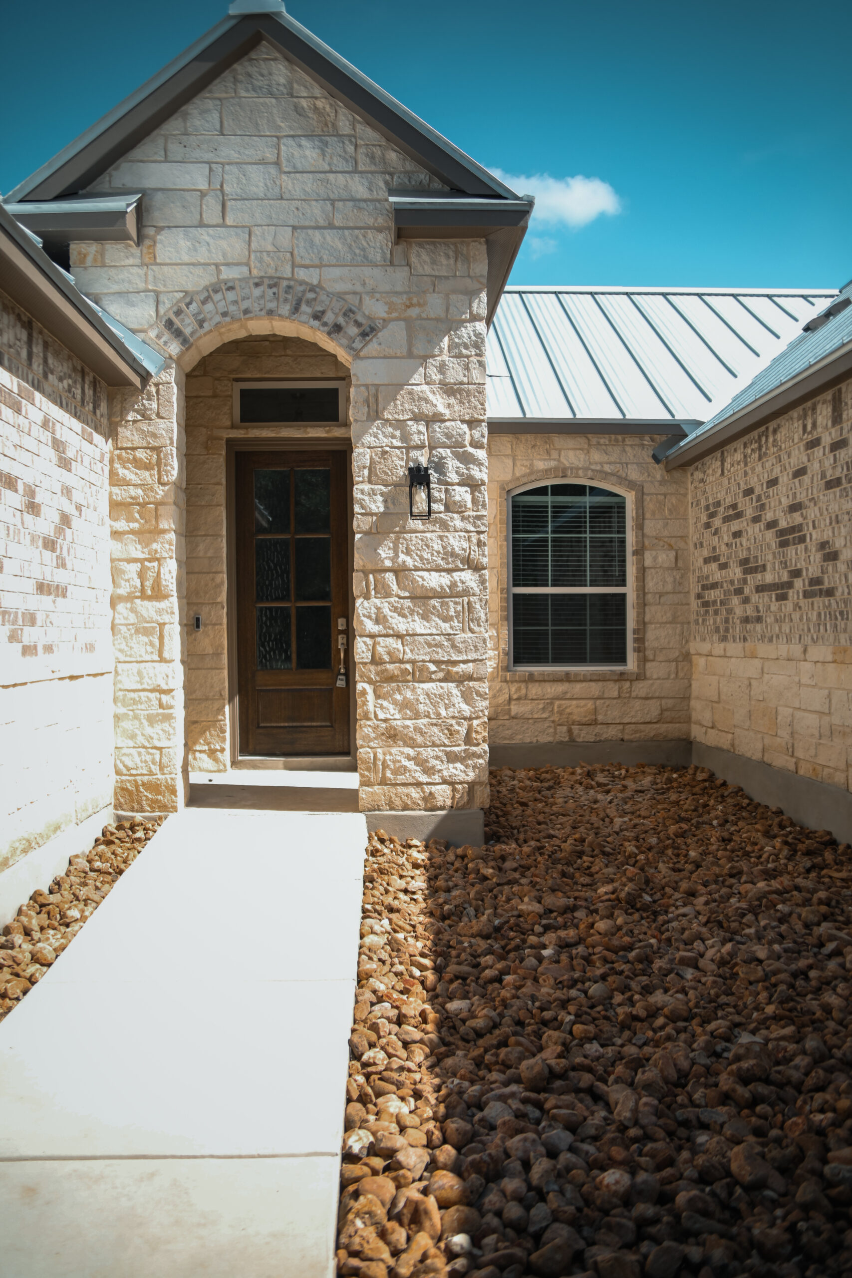A stone walkway leads to a wooden front door of a light-colored stone house with a metal roof and an arched entryway.