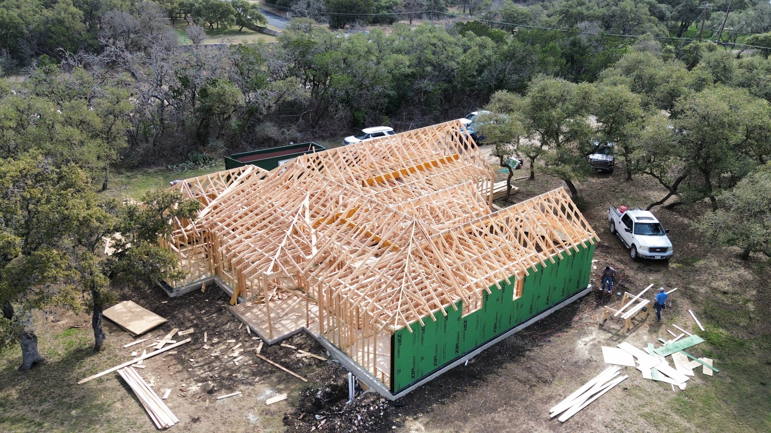 Aerial view of a house under construction with wooden framing and green sheathing, surrounded by trees and several parked vehicles.
