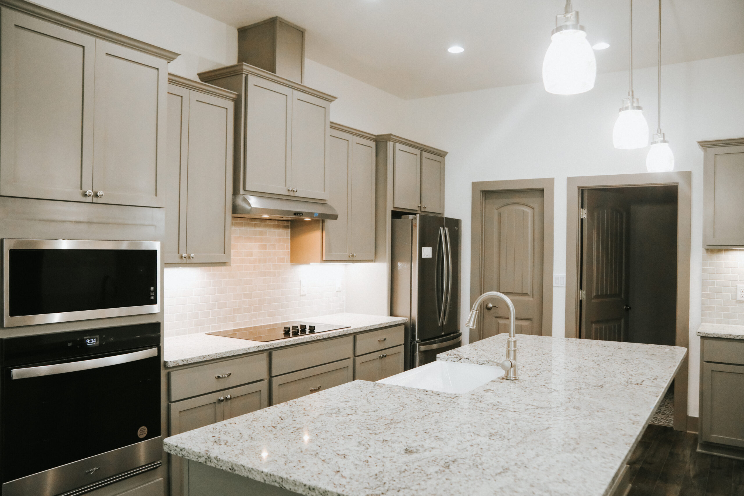 Modern kitchen with gray cabinets, stainless steel appliances, a large granite island with a sink, and pendant lighting.