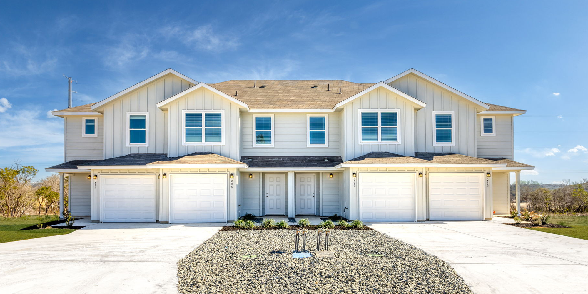 A modern two-story duplex with white siding, four garage doors, and a shared driveway, set against a blue sky.