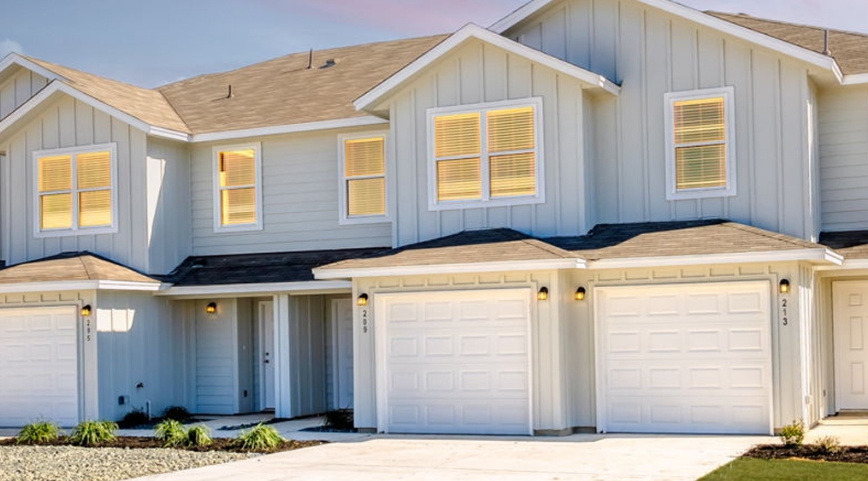 Two-story, light gray duplex homes with white trim, each featuring a single-car garage, double windows, and a small driveway.