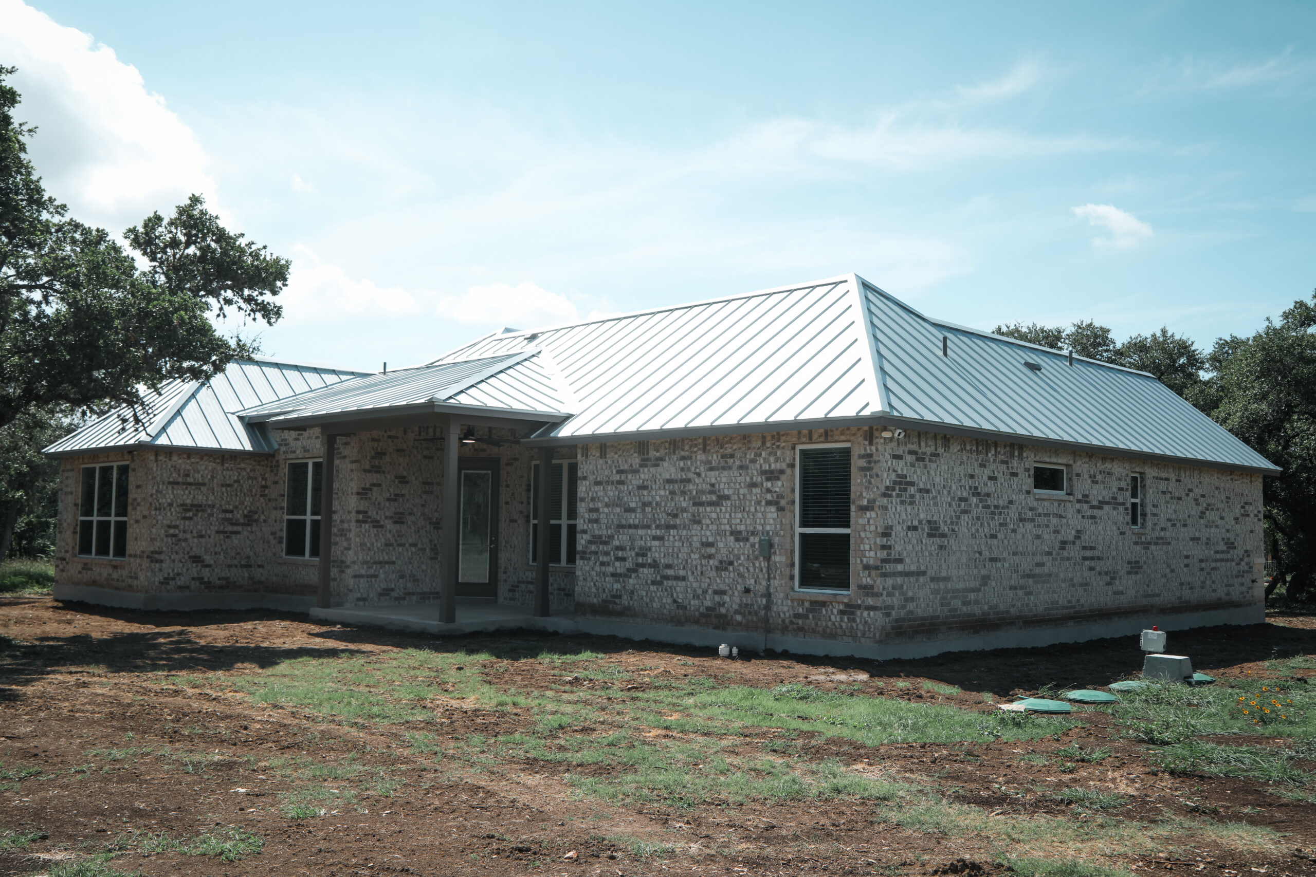 Single-story brick house with a metal roof, surrounded by bare ground and a few trees under a clear sky.