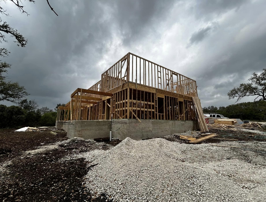 A two-story wooden house frame under construction stands on a concrete foundation, with building materials scattered around and a cloudy sky overhead.